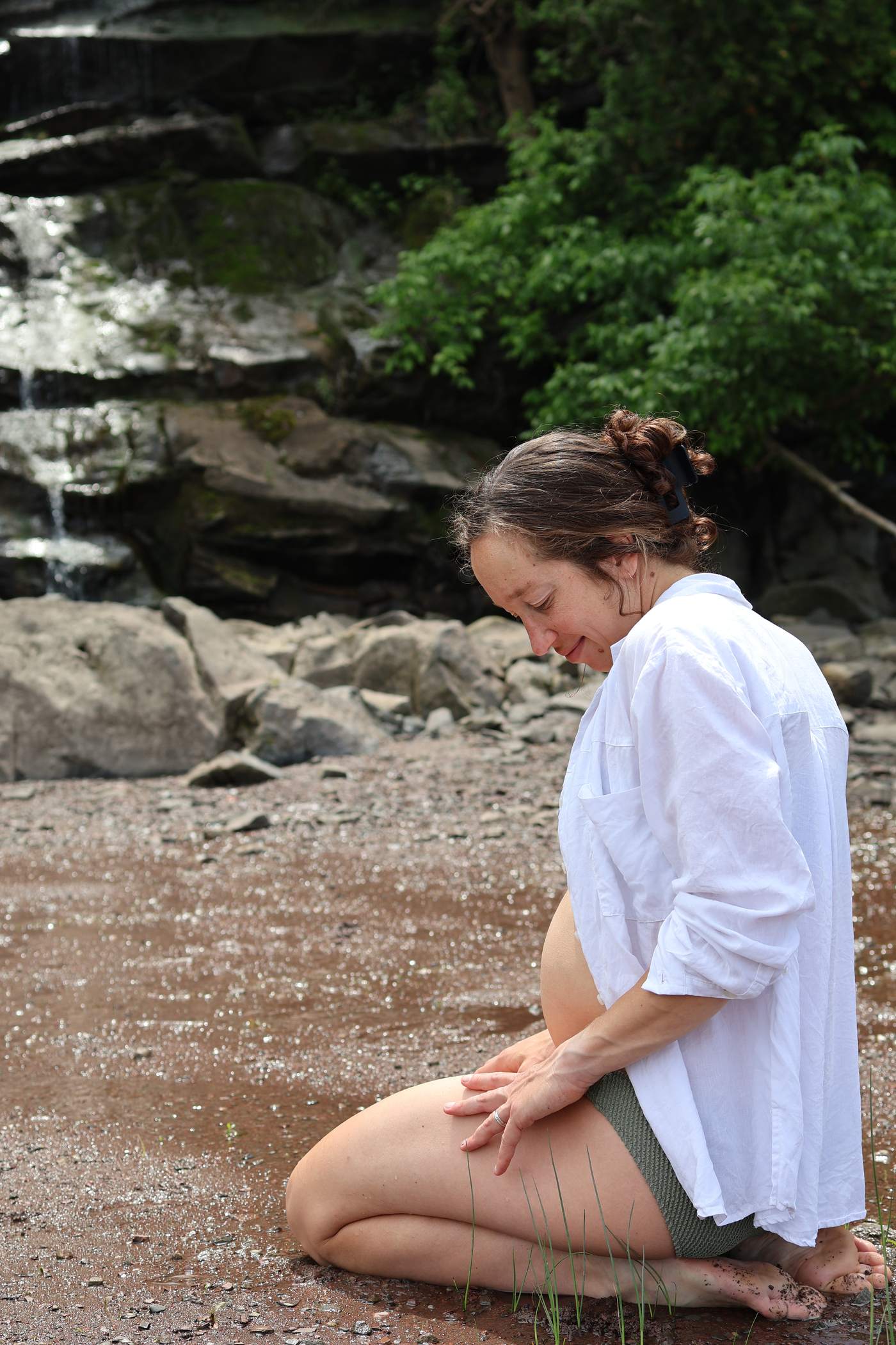 Femme enceinte agenouillée sur la grève en chemise blanche, une cascade derrière elle