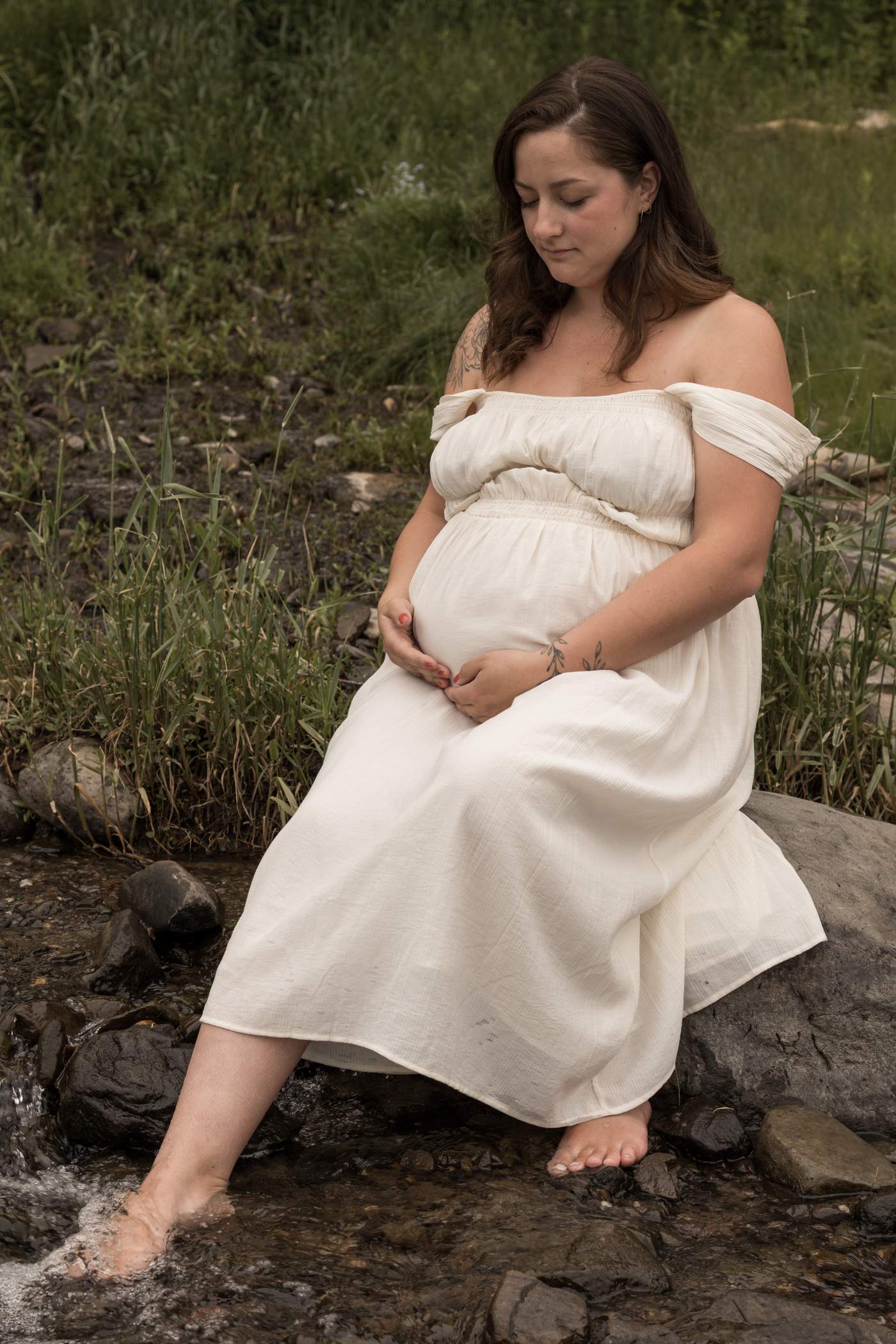 Femme enceinte en robe crème assise dans un ruisseau, photographiée par Anne-Sophie Fortier
