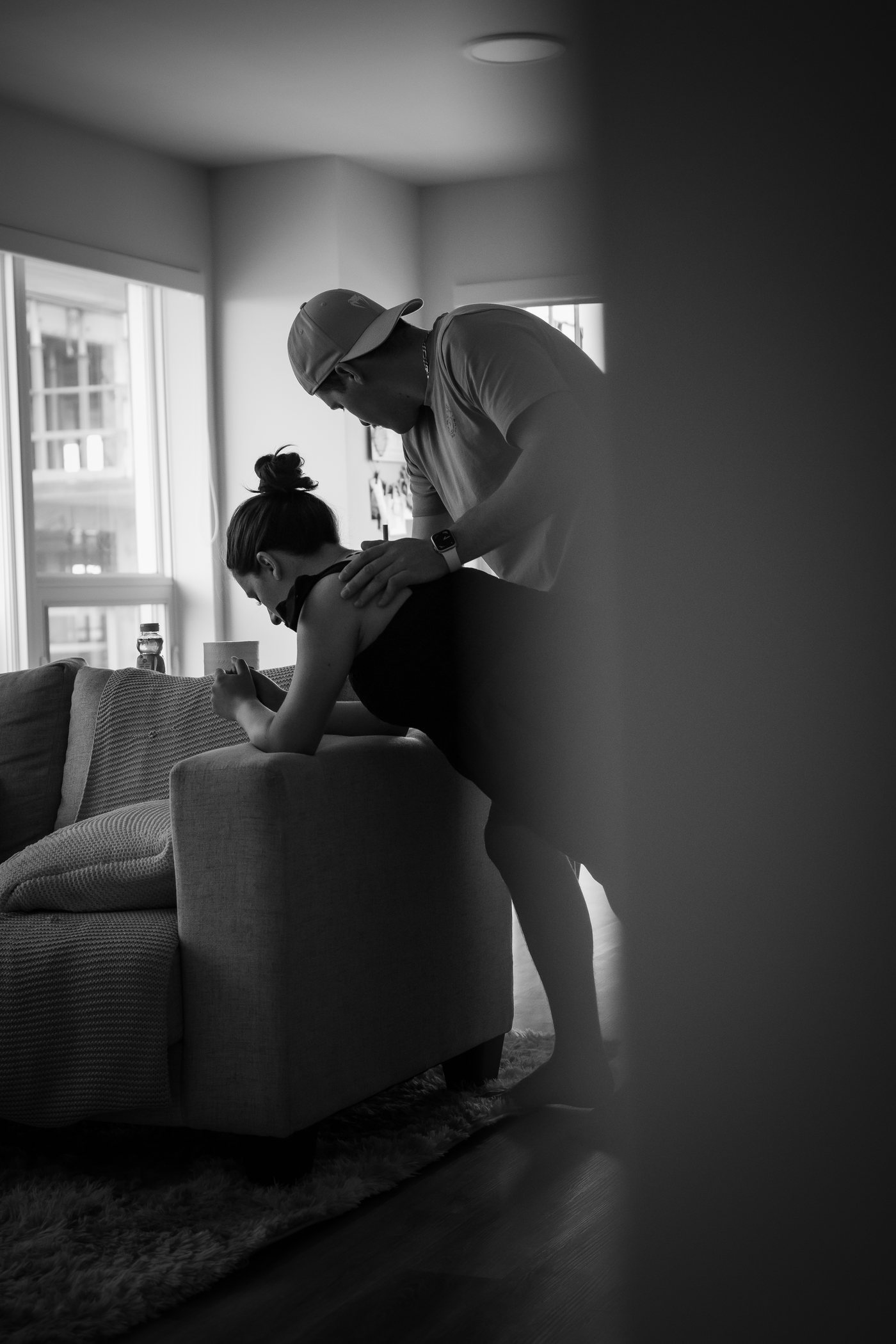 En noir et blanc, une femme en travail appuyée sur un divan, son partenaire penché sur elle